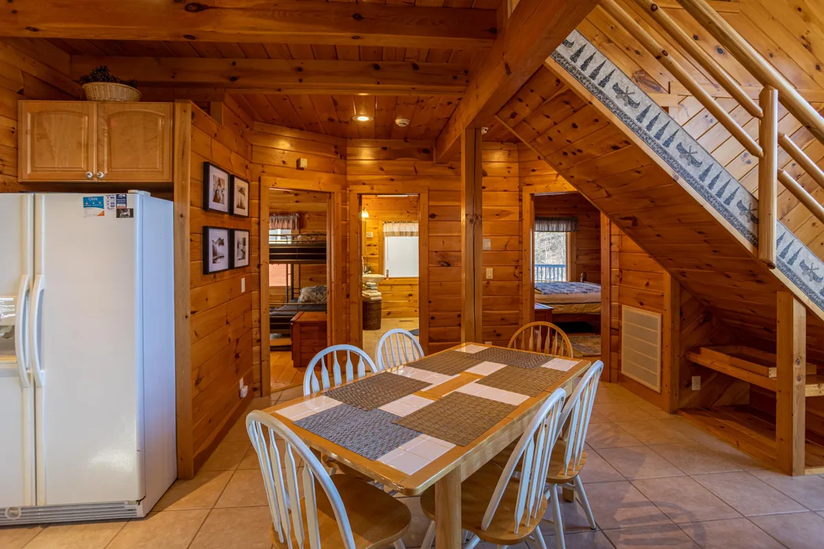 The dining area is in focus here, with a wooden table set for six, a staircase in the background, and wood accents throughout. The ambiance is homey and inviting, ideal for family meals, game nights, and storytelling long into the evening.