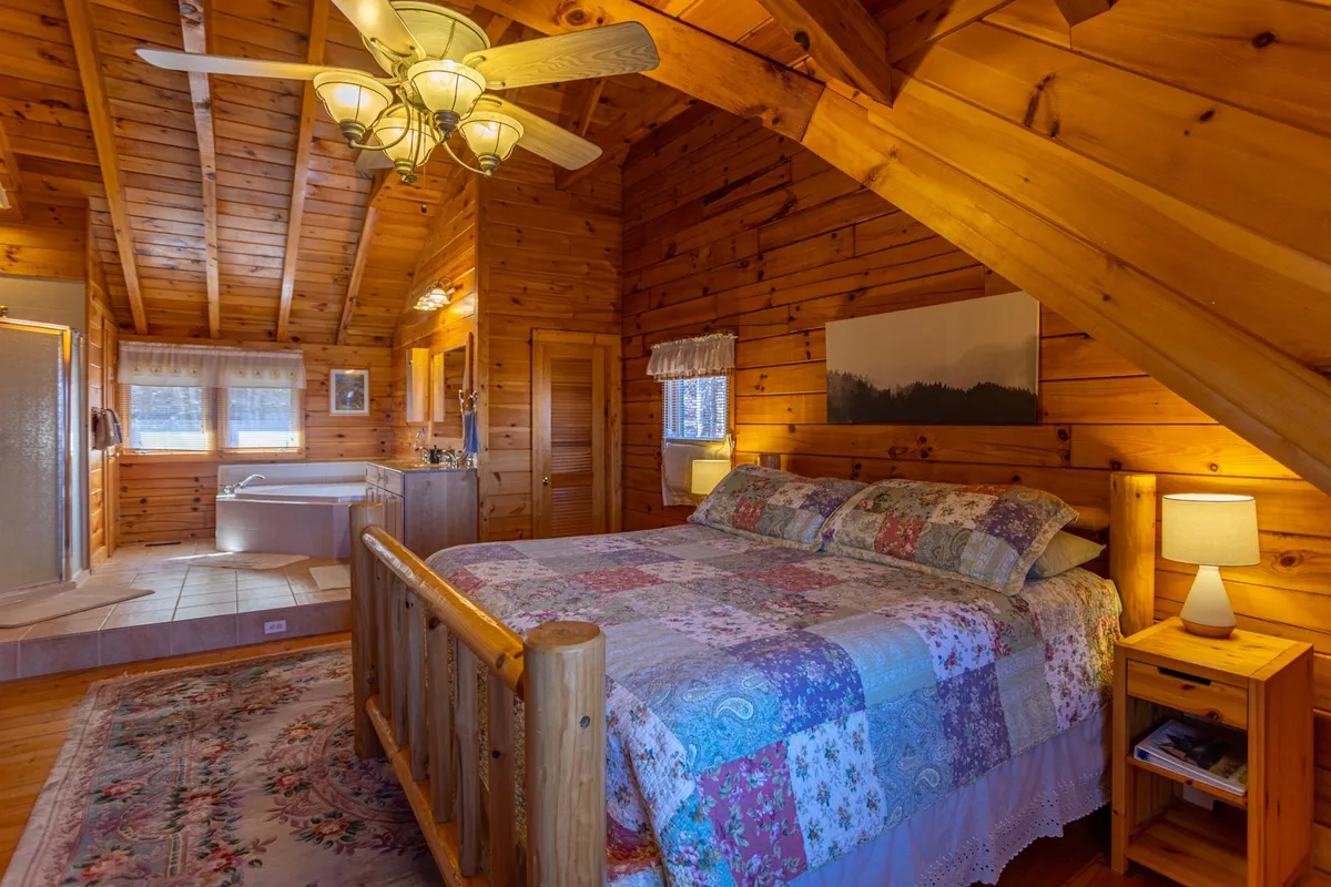 A cozy bedroom featuring a bed with a patchwork quilt, warm wooden walls and ceiling, and a jacuzzi tub nestled in the corner. Soft lighting from bedside lamps creates a welcoming atmosphere, and the room exudes comfort and tranquility—ideal for restful nights or a relaxing soak.