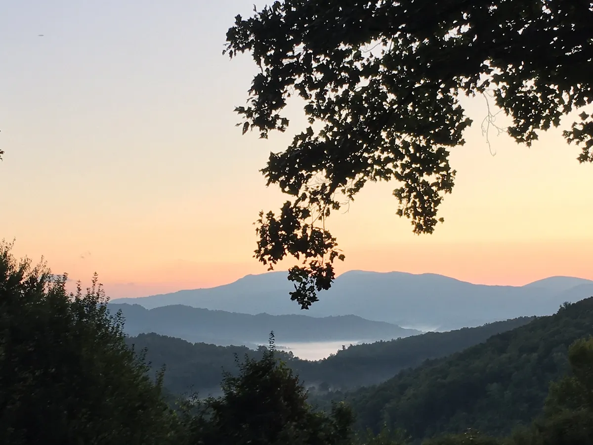 A breathtaking vista of distant mountains with fog drifting through the valleys and the warm glow of a sunset illuminating the landscape. The scene invites a sense of tranquility and connection to the surrounding wilderness, perfectly capturing the allure of the high country near Boone.