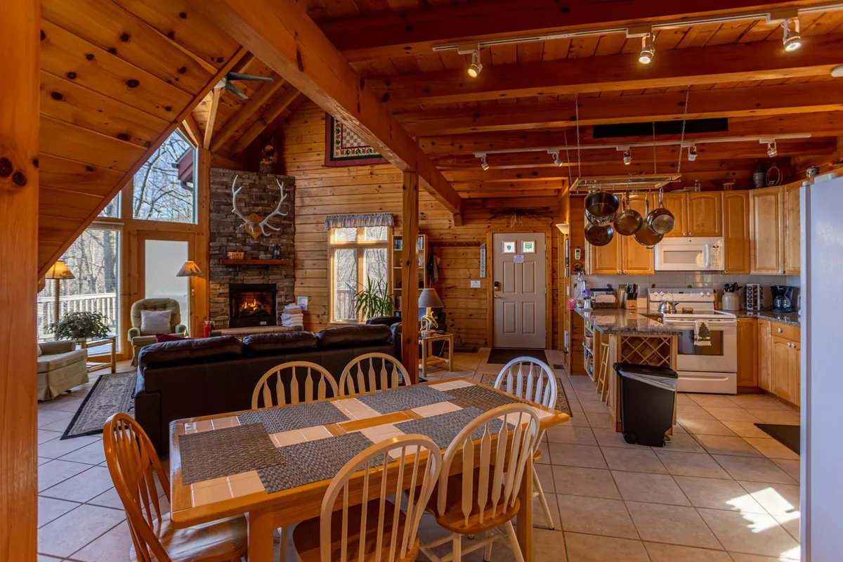 A wide shot shows the open-concept living and dining area, seamlessly connecting a welcoming stone fireplace, a large dining table, and the kitchen beyond. Wooden beams overhead and soft lighting create a heartwarming atmosphere, perfect for family gatherings or holiday celebrations.