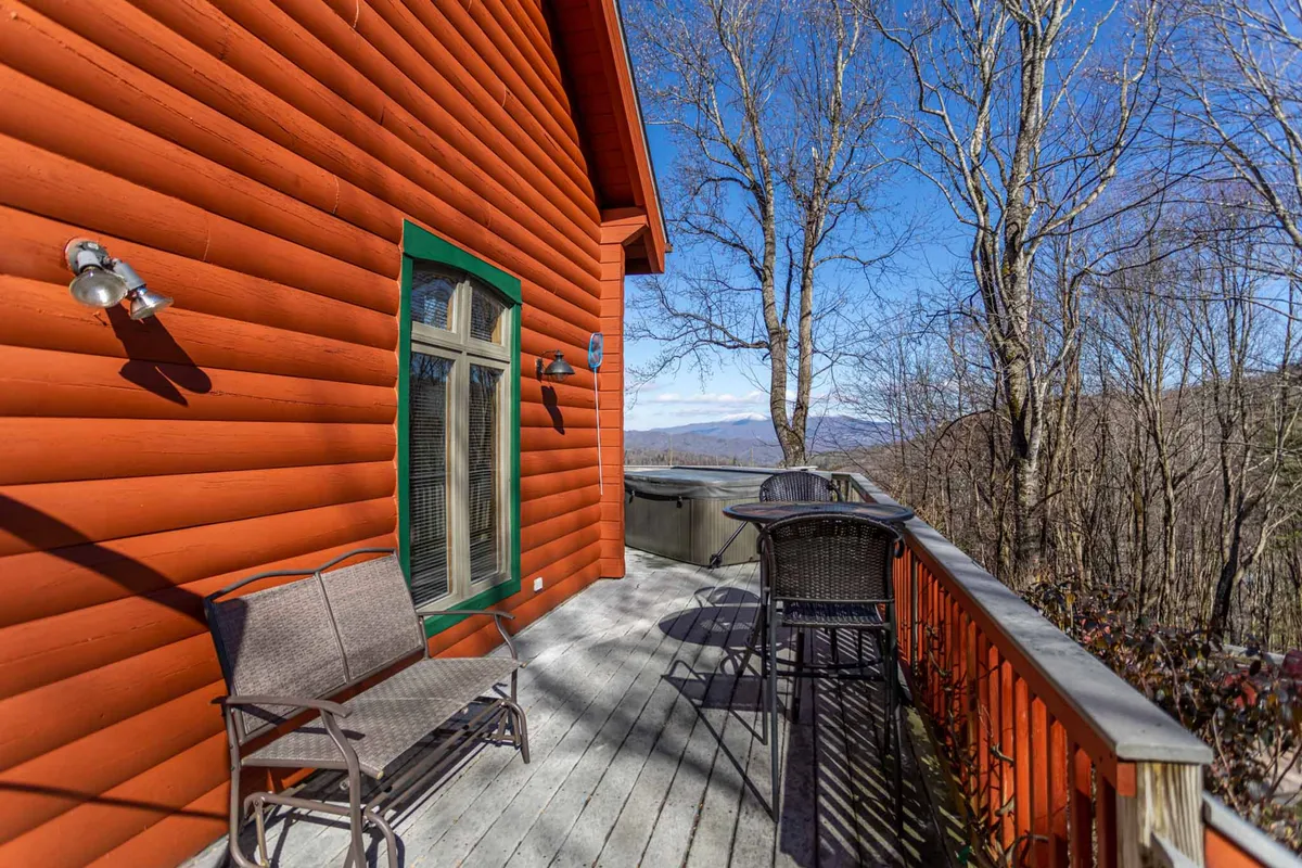 This exterior shot highlights the cabin's vibrant orange log siding and green window frames, with comfortable outdoor seating available on the deck. The setting is tranquil, inviting guests to relax, socialize, or stargaze while enjoying the mountain views.