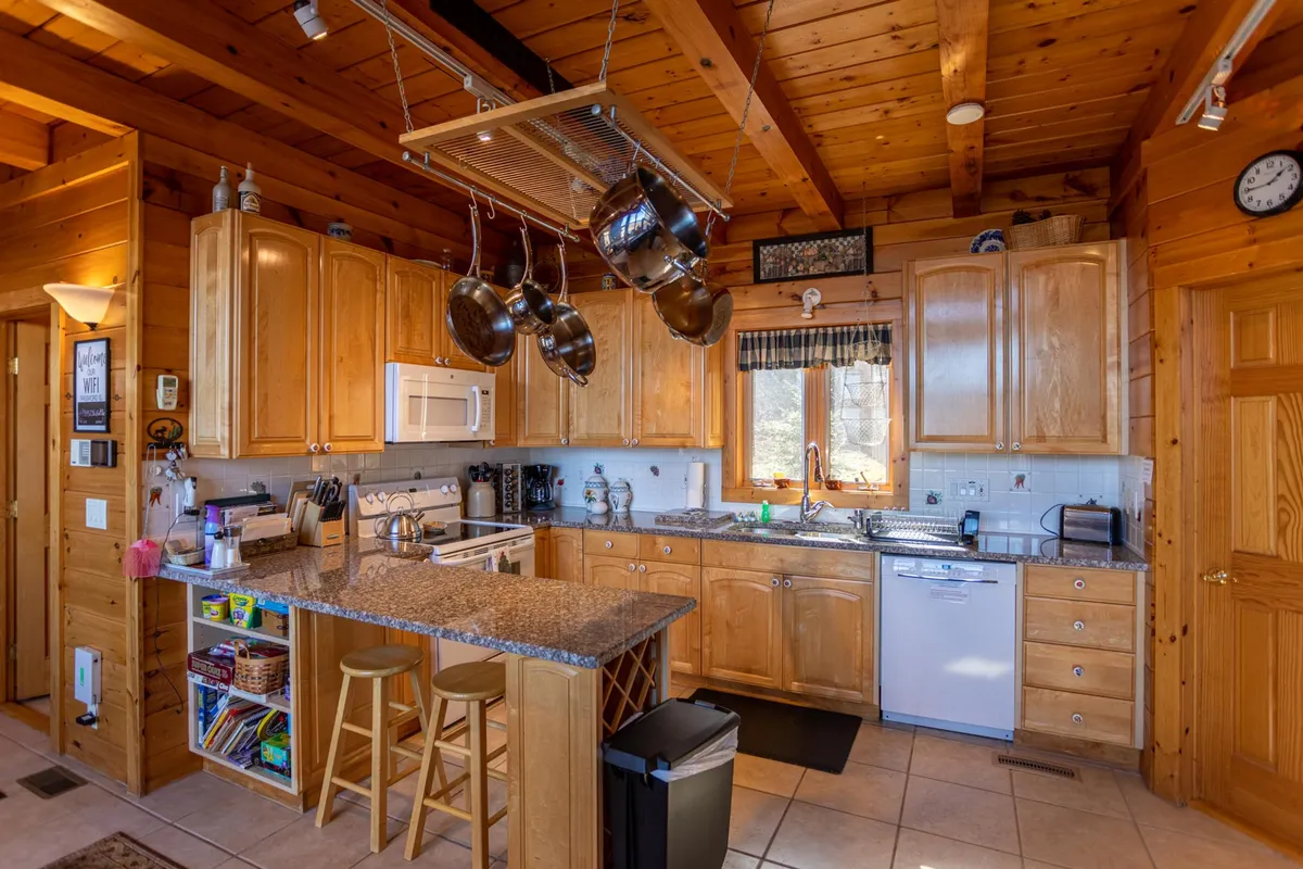 A warm, inviting kitchen scene featuring wooden cabinetry, granite countertops, hanging pots and pans, and a window that offers a scenic view. Bar stools line the counter, ready for casual dining or socializing. The space appears stocked and ready for culinary adventures, with clear sight lines that encourage interaction.