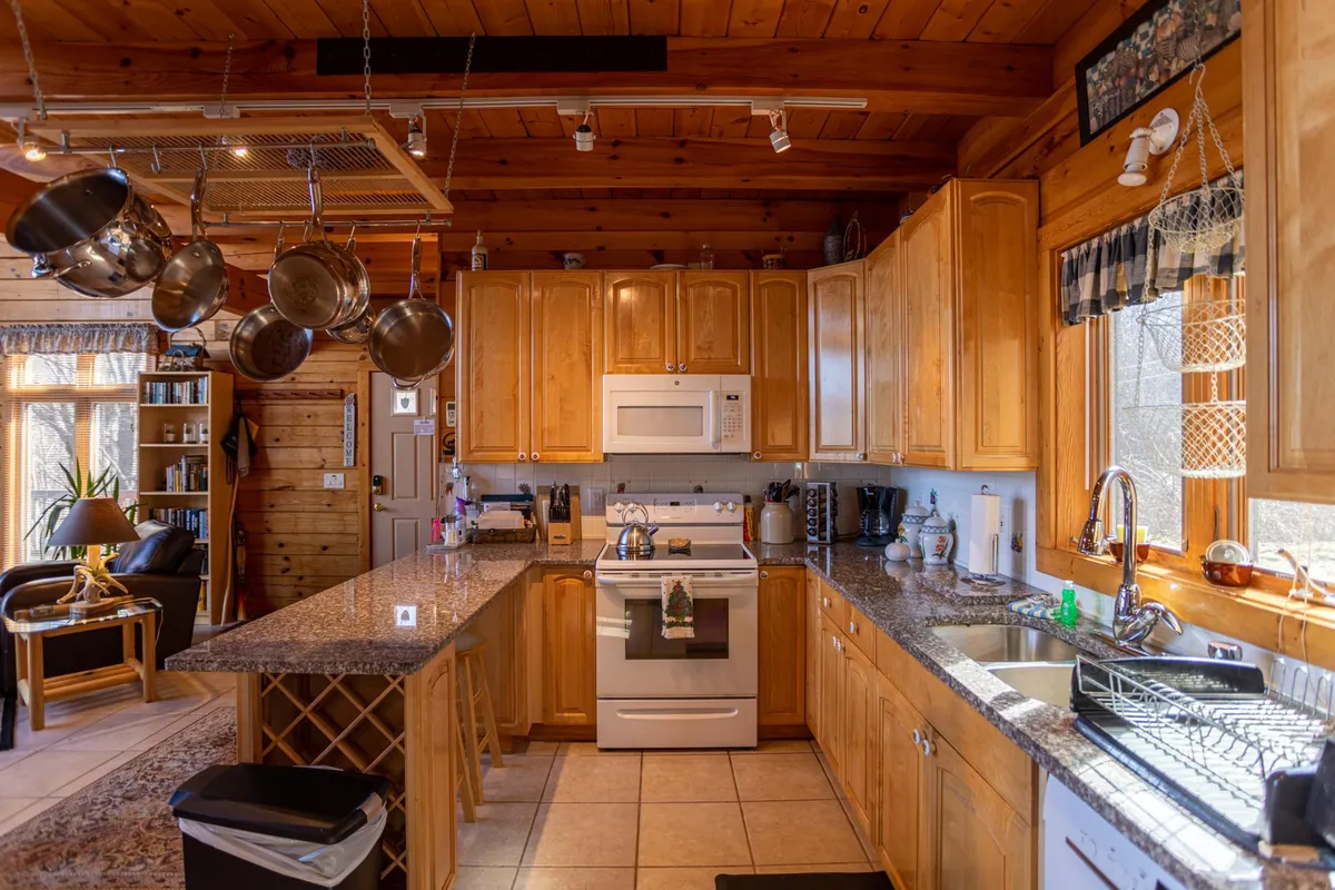 A cozy kitchen with a granite-topped island, wooden cabinetry, large windows letting in natural light, and hanging pots above the workspace. The arrangement encourages both cooking and gathering, with ample counter space for meal prep or coffee chats.