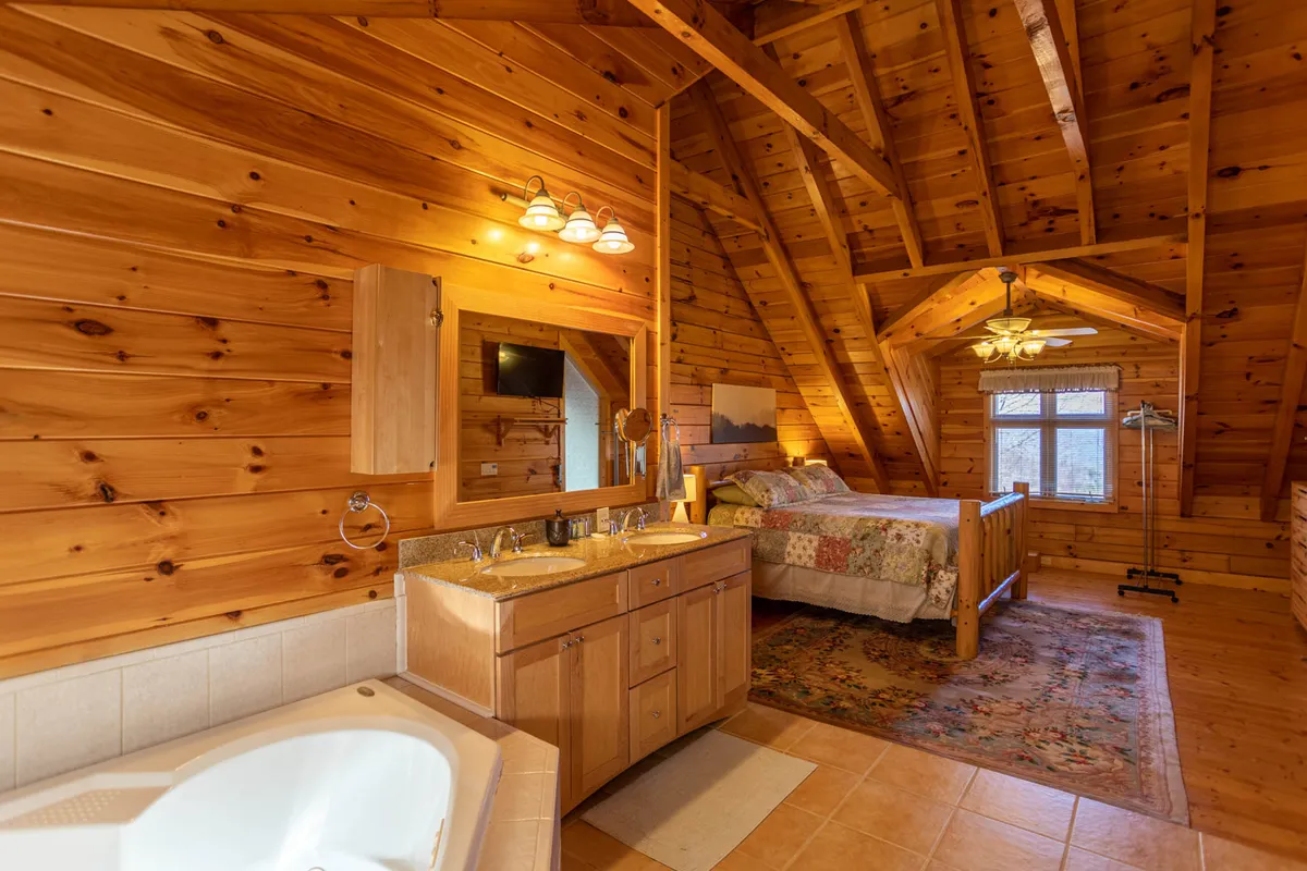A serene, open-plan master suite featuring exposed wooden beams, a comfortable bed with a quilt, a large window letting in natural light, and a bathtub located within view of the outdoors. The space feels luxurious, airy, and uniquely designed for romance and relaxation.