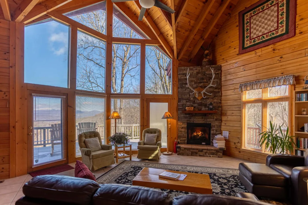 This image features the living room with comfortable seating arranged around a stunning rustic stone gas fireplace. Large windows allow natural light to flood the space, while wooden beams and a decorative quilt add warmth and charm. The scene captures guests relaxing by the fireplace, reading, or enjoying the view outside.
