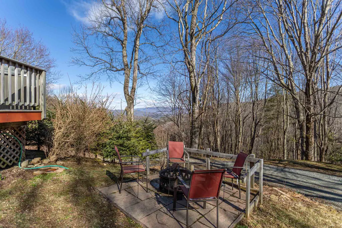 A cozy outdoor fire pit area set against a stunning mountain backdrop, surrounded by trees and comfortable patio seating. The scene evokes evenings spent gathering, sharing stories, and enjoying the mountain air, with sunset light creating a warm, inviting mood.