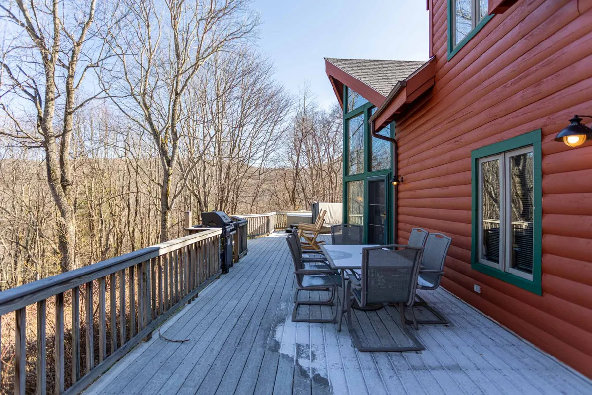 This image shows a welcoming outdoor deck area complete with a table, chairs, grill, and large windows looking into the cabin. The scene is set for outdoor dining, barbecuing, and relaxing, surrounded by the natural mountain environment.
