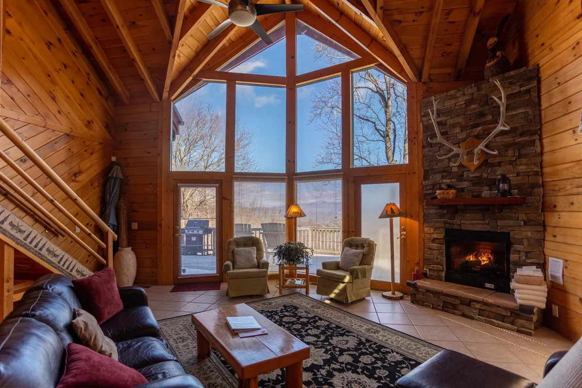 The image captures the cozy cabin interior with soaring wooden beams, a crackling stone fireplace, plush seating, decorative antlers, and expansive windows framing mountain views. The space feels inviting and perfect for gathering, unwinding by the fire, or soaking in the natural beauty outside.